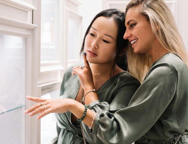 Two women from a bridal party wearing matching outfits in a jeweler’s showroom after finding them online through keyword searches to explore beautiful jewelry options for the bridesmaids and bride.