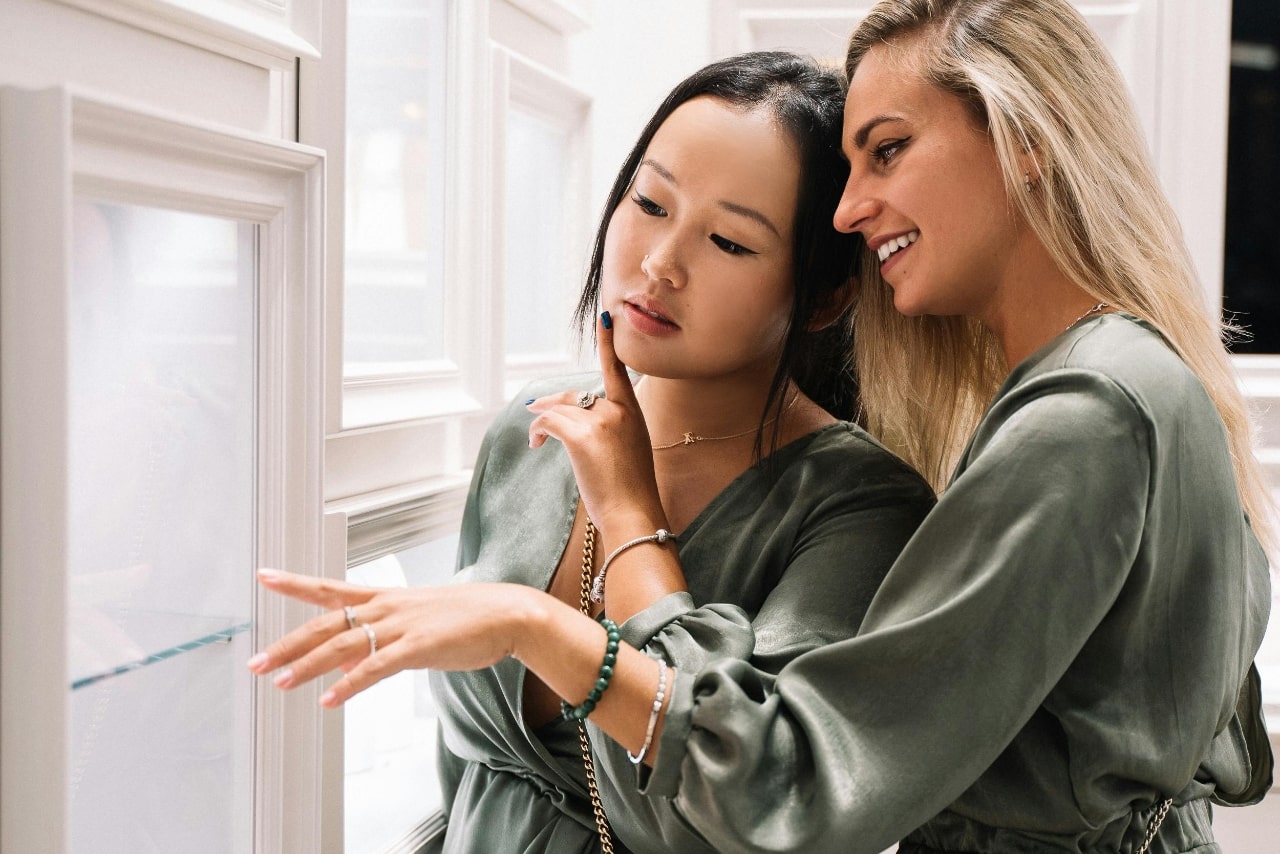 Two women from a bridal party wearing matching outfits in a jeweler’s showroom after finding them online through keyword searches to explore beautiful jewelry options for the bridesmaids and bride.